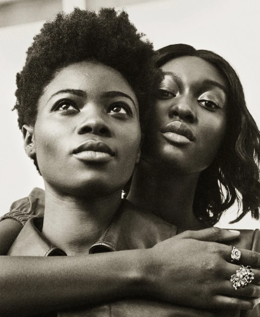 Monochrome portrait of two Black women embracing, adorned with statement rings, in a bright indoor space. Fashion and beauty.