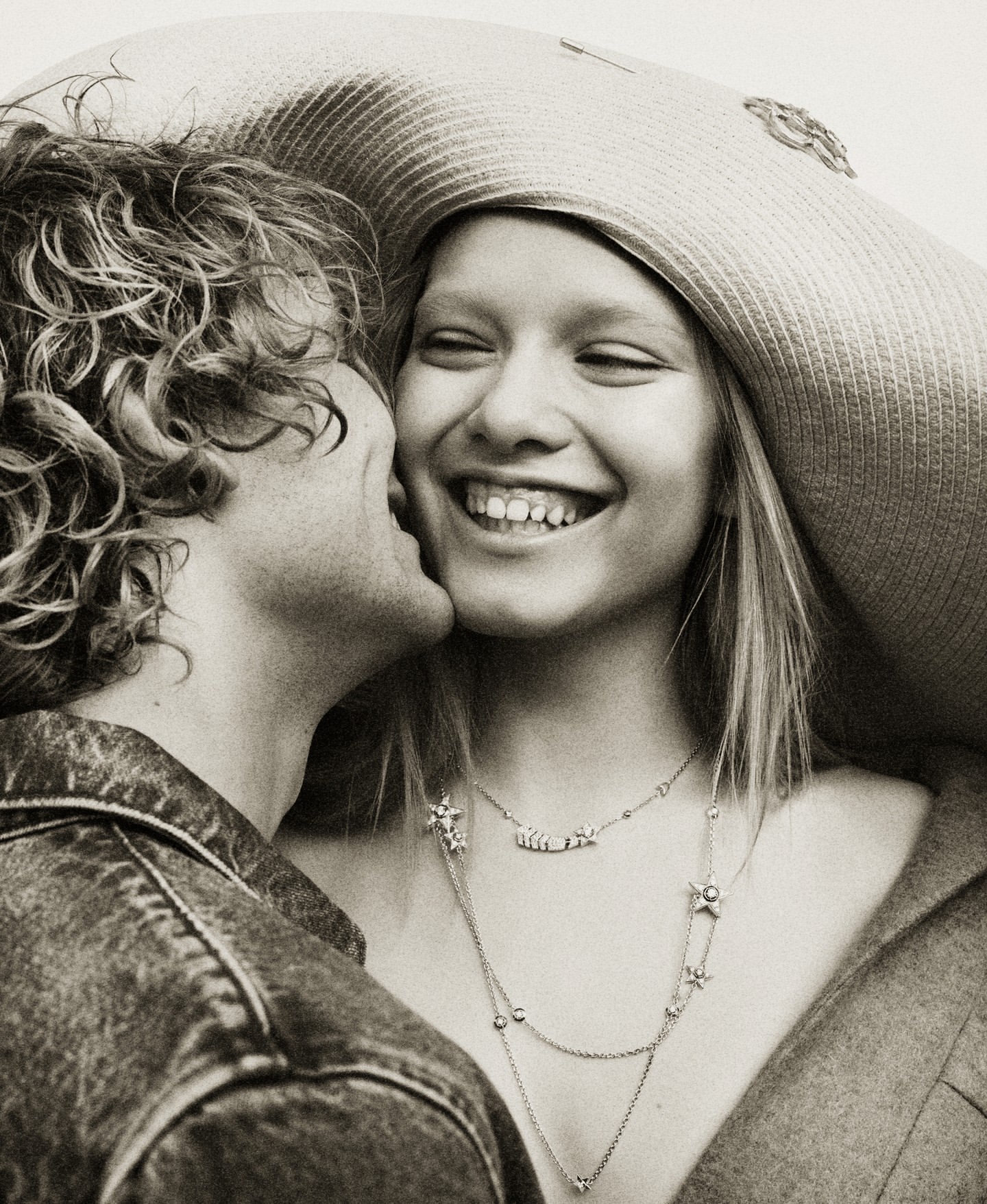 Romantic portrait of a smiling young woman in a wide-brimmed hat receiving a kiss on the cheek from a curly-haired young man.
