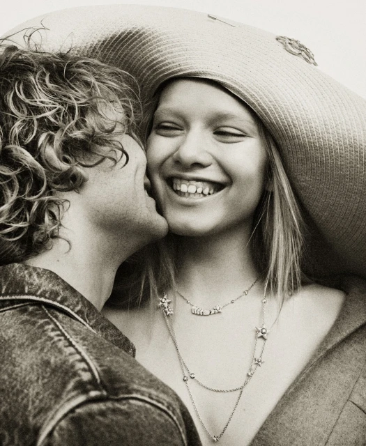 Romantic portrait of a smiling young woman in a wide-brimmed hat receiving a kiss on the cheek from a curly-haired young man.