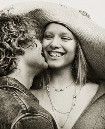 Romantic portrait of a smiling young woman in a wide-brimmed hat receiving a kiss on the cheek from a curly-haired young man.