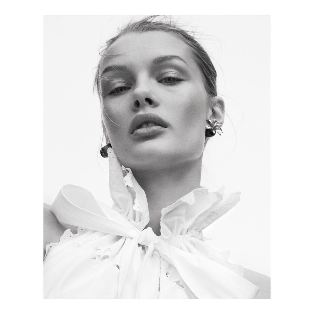Black and white portrait of a woman wearing a ruffled white top and dark earrings against a bright, neutral background, low angle.
