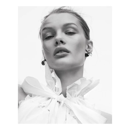 Black and white portrait of a woman wearing a ruffled white top and dark earrings against a bright, neutral background, low angle.