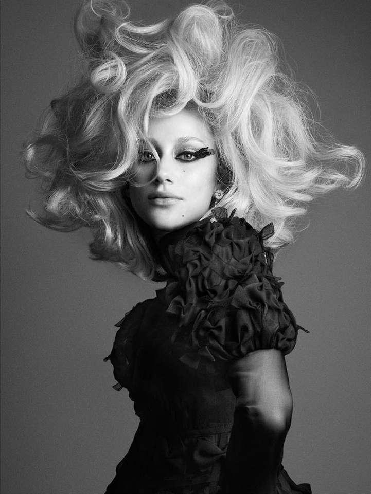 Black and white portrait of a fair-skinned woman with teased blonde hair, heavy eye makeup, and a black ruffled dress against a grey background.