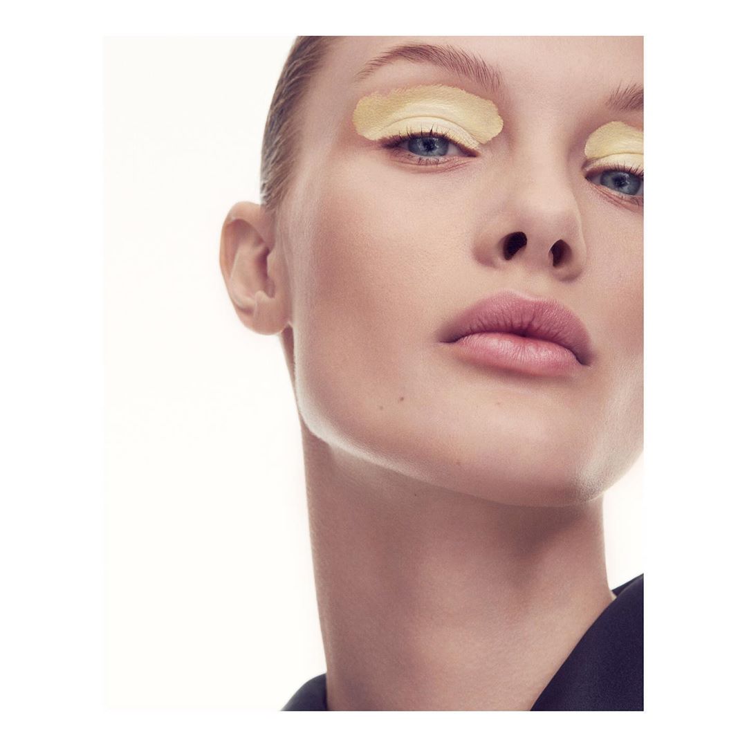 Close-up studio shot of an adult female with light skin, blonde hair, and yellow eye makeup, wearing a black top against a white backdrop.