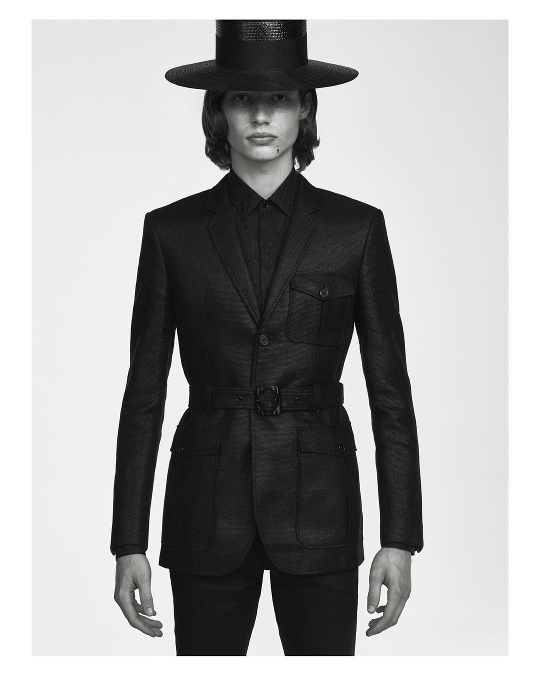 Monochromatic portrait of a slender man in a dark suit, hat, and belt, set against a neutral background with soft, diffused lighting.