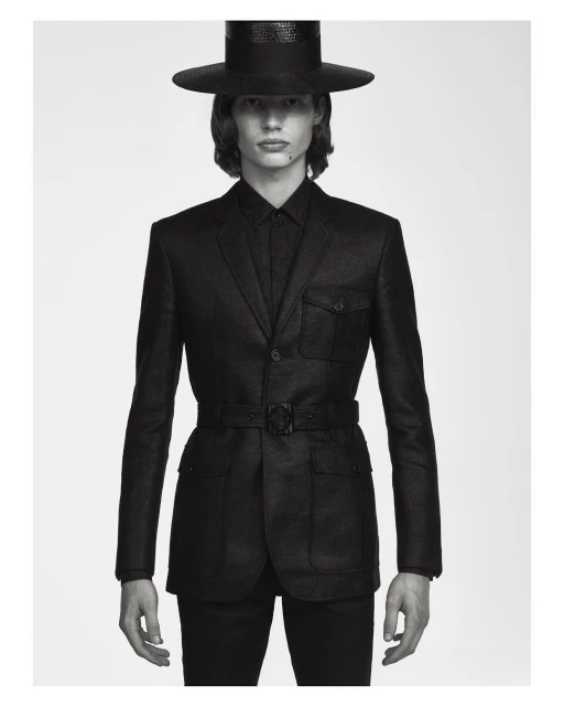 Monochromatic portrait of a slender man in a dark suit, hat, and belt, set against a neutral background with soft, diffused lighting.