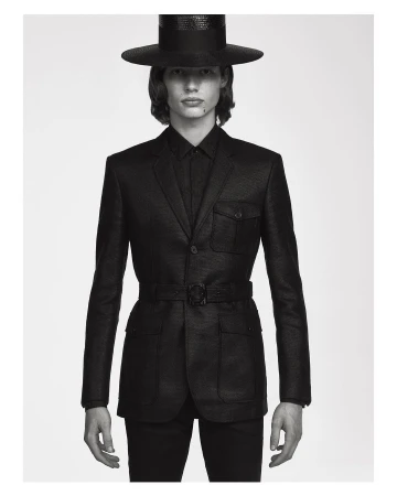 Monochromatic portrait of a slender man in a dark suit, hat, and belt, set against a neutral background with soft, diffused lighting.