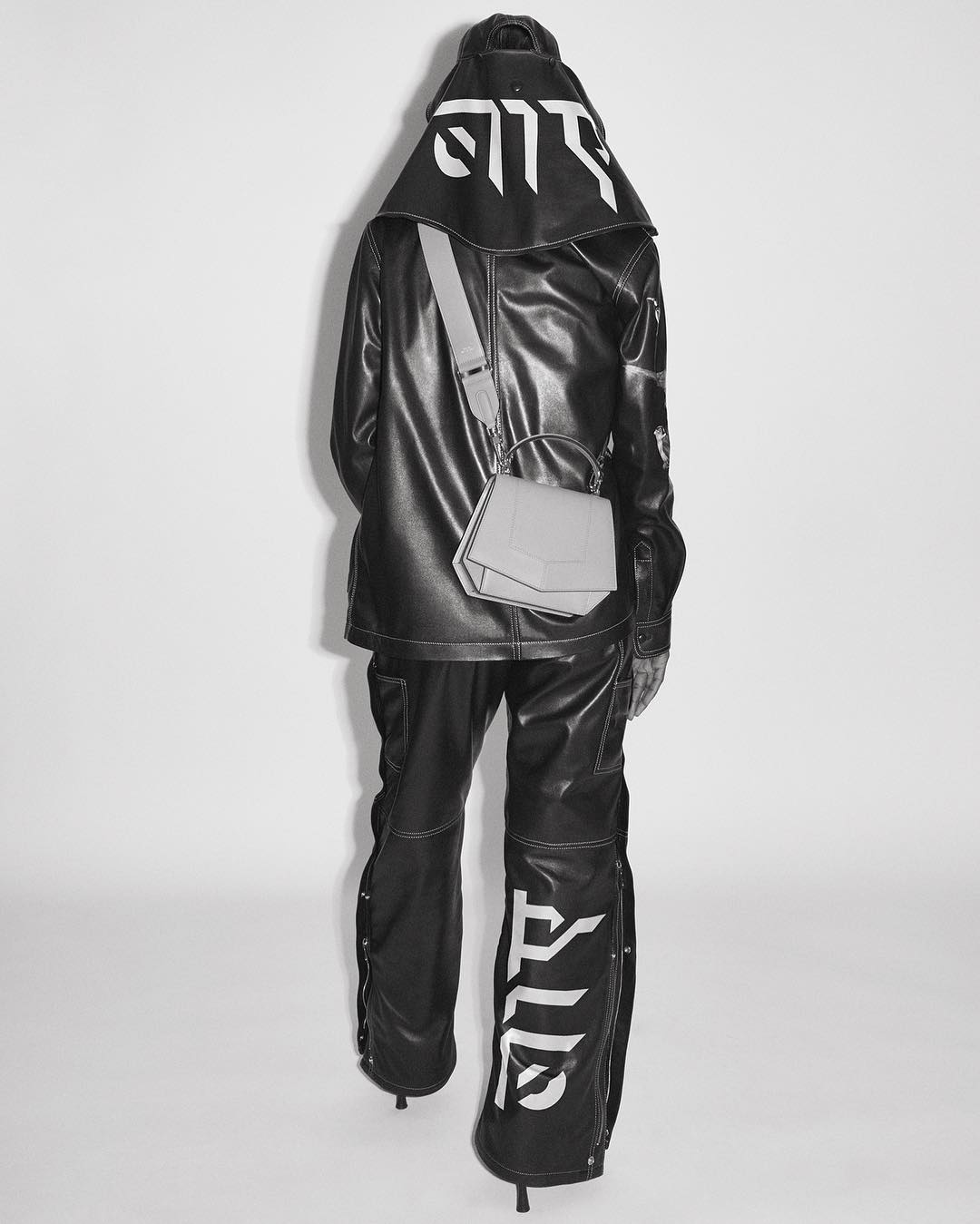 Fashionable person in black vinyl hooded jacket and pants with white graphics, accessorized with a geometric shoulder bag, standing on white.