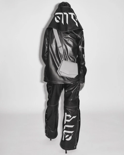 Fashionable person in black vinyl hooded jacket and pants with white graphics, accessorized with a geometric shoulder bag, standing on white.