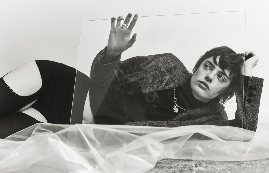 Monochrome portrait of a woman in a blazer and stockings, striking a pose with an elevated hand against a clear plastic sheet.