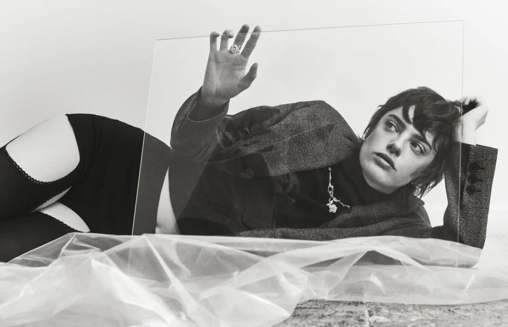 Monochrome portrait of a woman in a blazer and stockings, striking a pose with an elevated hand against a clear plastic sheet.