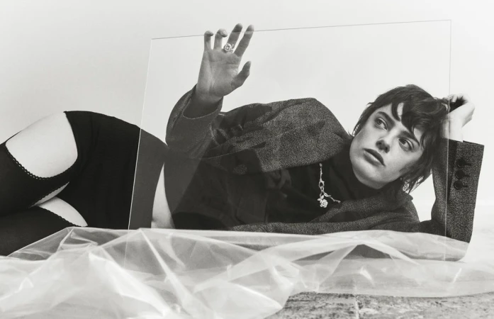 Monochrome portrait of a woman in a blazer and stockings, striking a pose with an elevated hand against a clear plastic sheet.