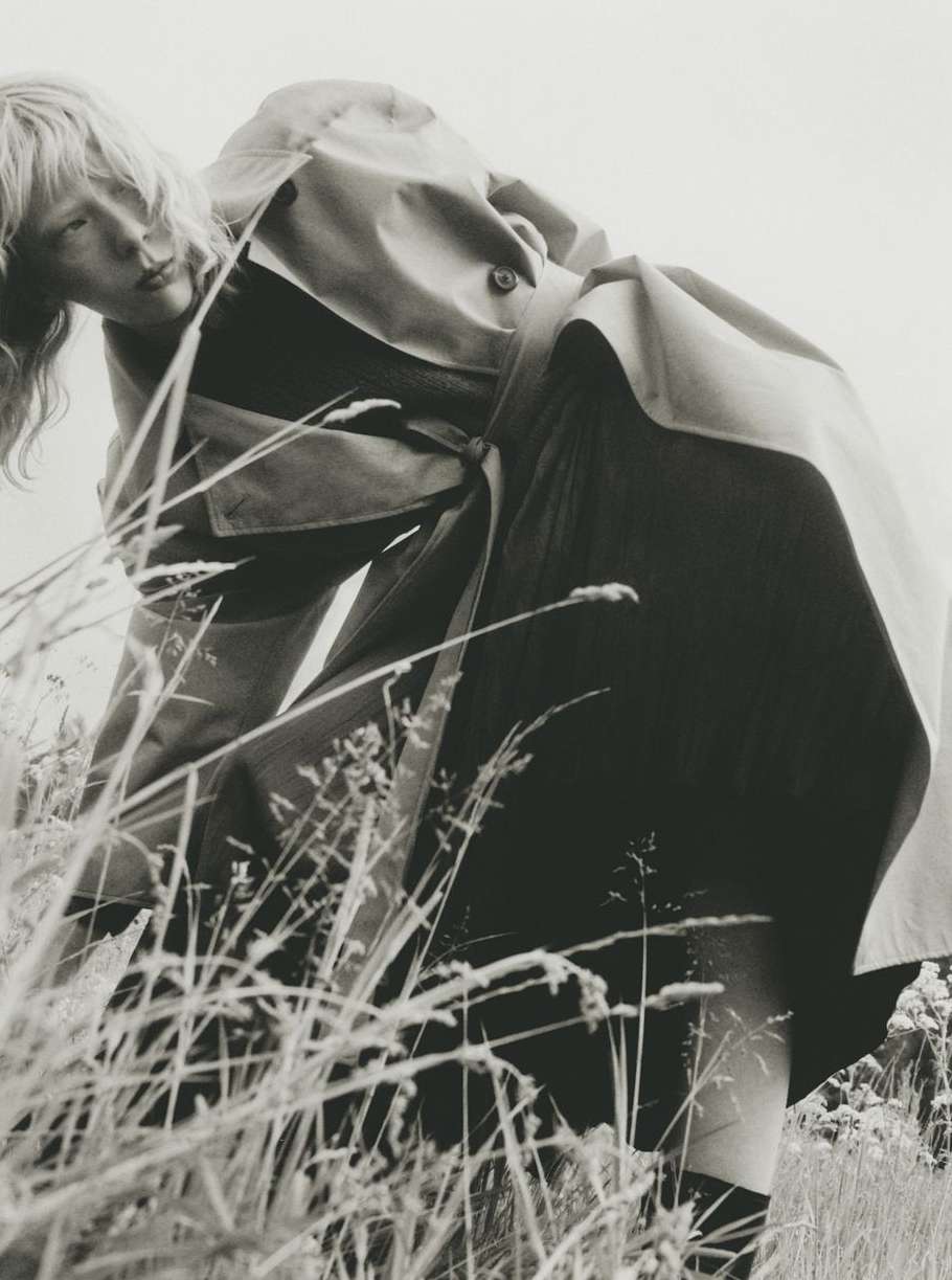 Model in trench coat leaning over tall meadow grass in a black and white editorial photograph by Marcus Ohlsson