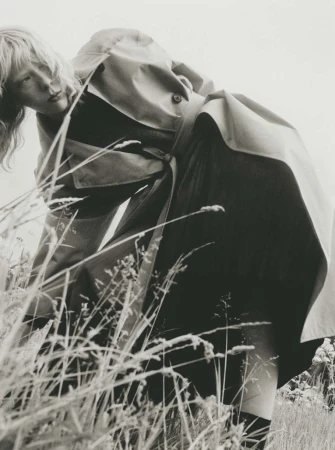 Model in trench coat leaning over tall meadow grass in a black and white editorial photograph by Marcus Ohlsson