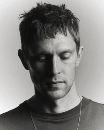 Close-up monochrome portrait of a man with short dark hair and a thin silver necklace against a plain grey background