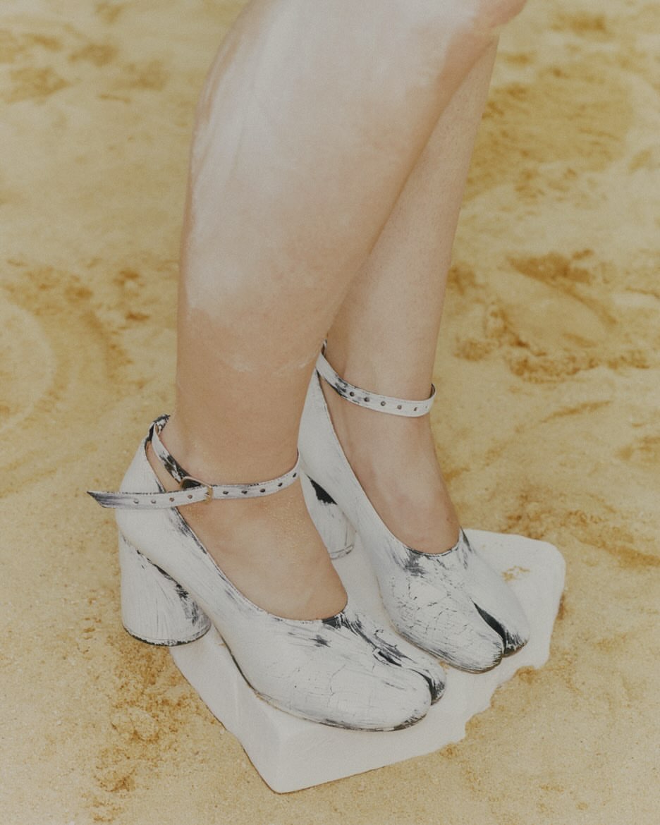 A person models painted Tabi heels with ankle straps against a textured tan backdrop, legs partially visible in this fashion-focused shot.