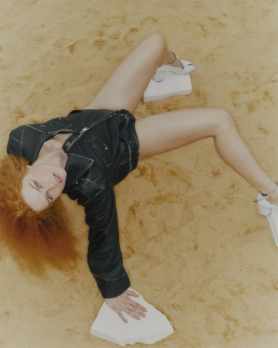 An adult woman with red hair lies on a sandy surface, wearing a dark jacket and shorts, and posing with white rectangular objects.