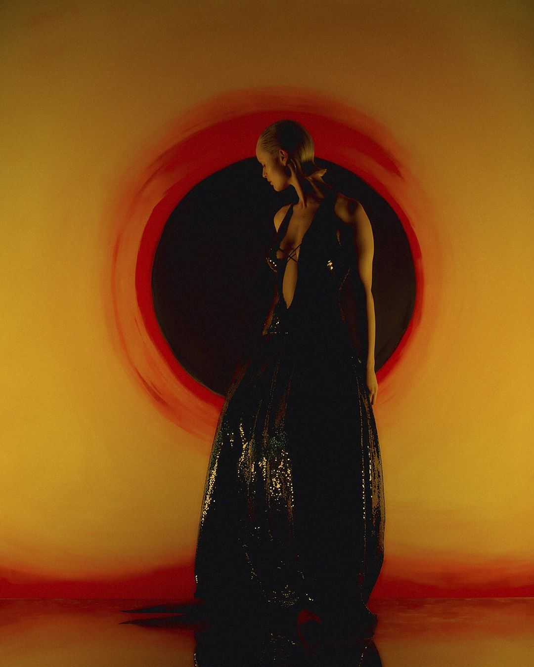 Back view of a female adult wearing a black sequined gown standing indoors in front of a red and orange round painted background.