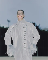 Model wearing a light grey high-neck coat with feathered cuffs and a dotted face veil outdoors by Marek Micanek