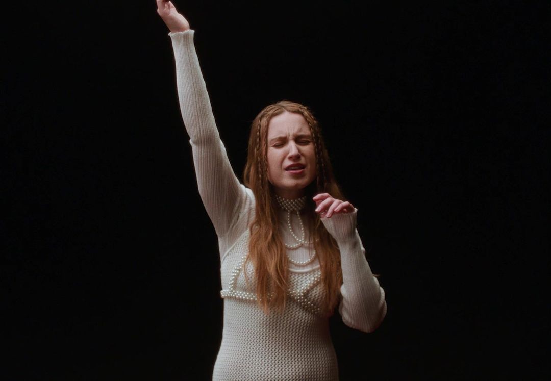Woman in a pearl-covered dress performs with arm raised against a stark black backdrop, creating a dramatic and captivating visual.