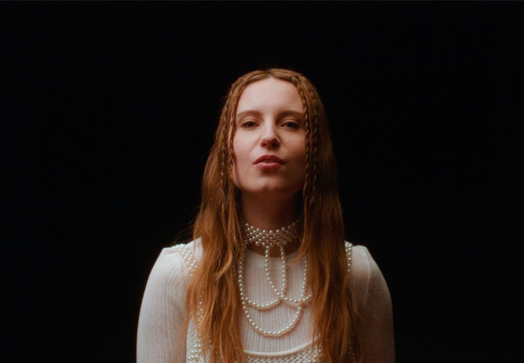 Headshot of a red-haired woman against a black background, wearing a pearl beaded collar and white textured top, gazing at the camera.