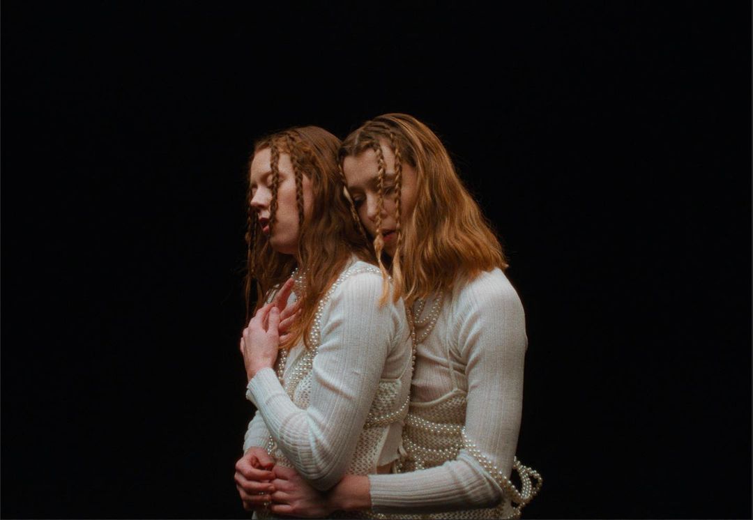 Two adult women embrace in a studio portrait against a dark background, wearing matching white sweaters and strands of pearls.