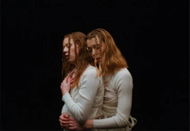 Two women with braided hair wearing white tops adorned with pearls embracing against a dark background