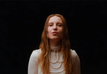 A woman with braided red hair wearing a white pearl-adorned top against a solid dark background.