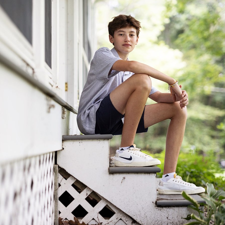 Teenager sits on white porch steps in casual clothes and sneakers with green leafy trees in the background. Summer casual fashion.