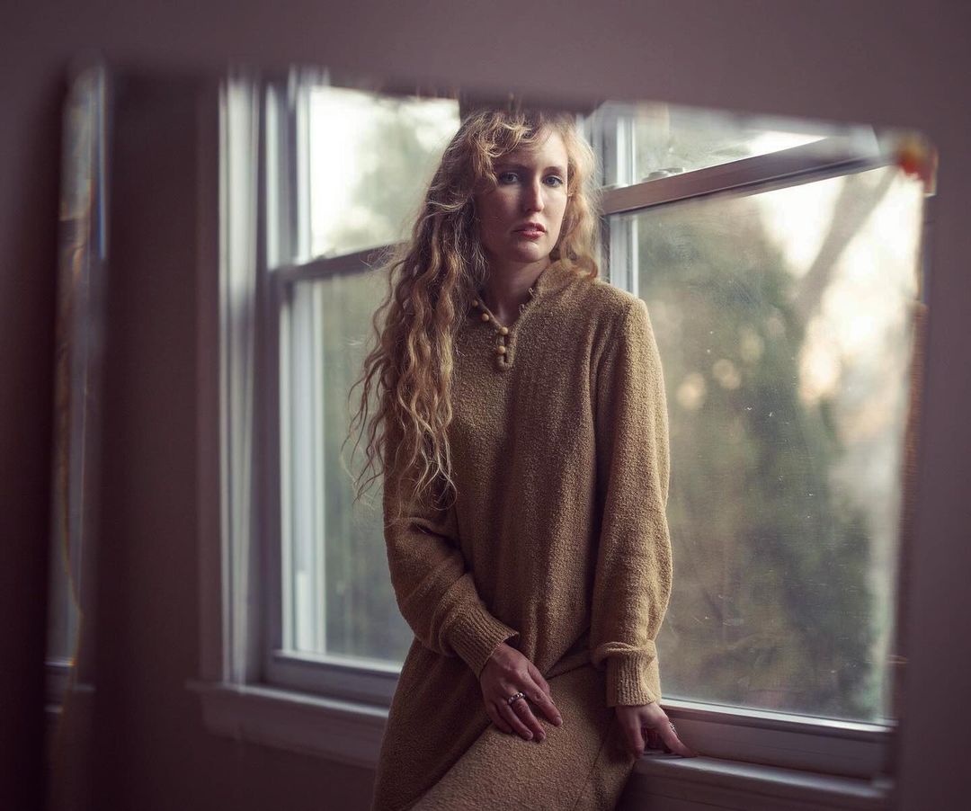 Portrait of a young woman with curly hair wearing a tan sweater looking through a window in a softly lit indoor setting.