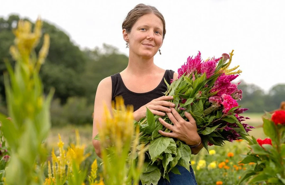 Woman holds large colorful flower bouquet in rural field with yellow, orange, red blooms under bright daylight.
