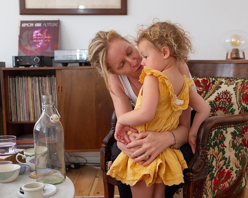 A mother tenderly holds her young child in a home interior setting, with vintage furniture and soft, natural lighting.