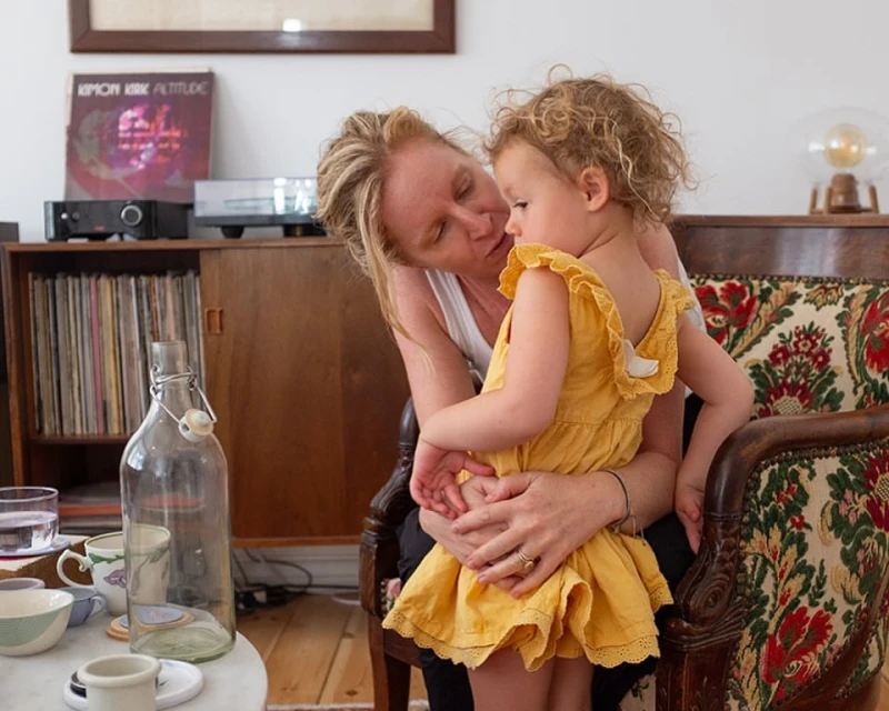 A mother tenderly holds her young child in a home interior setting, with vintage furniture and soft, natural lighting.