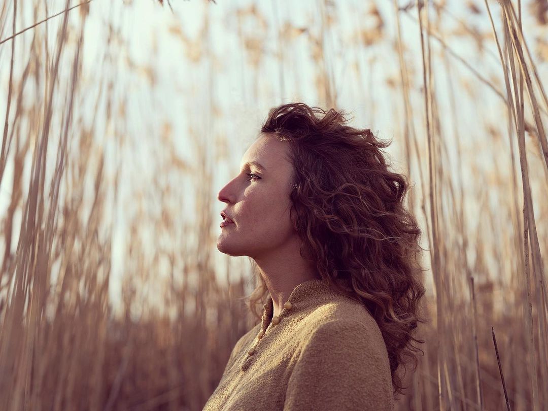 Woman with curly brown hair in profile stands surrounded by tall, dry reeds with a soft, golden light creating a serene natural scene.