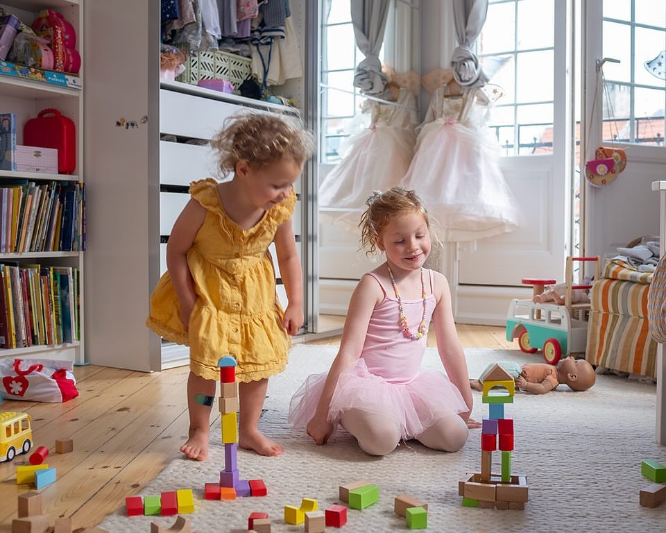 Two young girls are playing with colorful wooden blocks on a light rug in a brightly lit room, with wardrobe and children's toys visible.