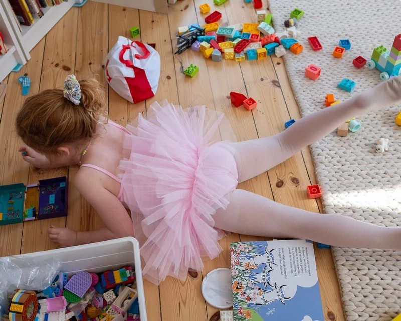 Young child in a pink tutu doing the splits on a wood floor surrounded by colorful building blocks and a book, indoors.