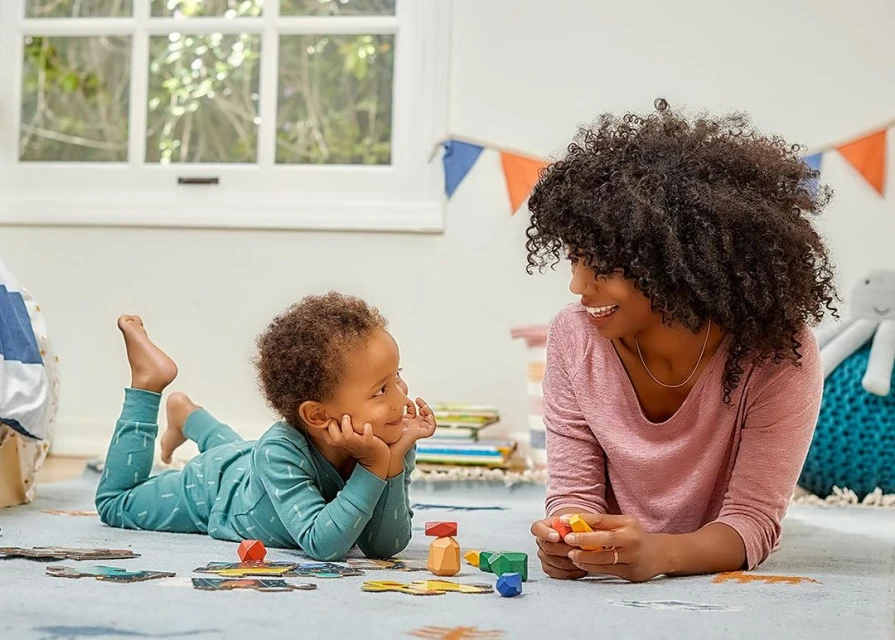 Young child lying on the floor playing a puzzle with an adult female who is smiling and looking at him inside a brightly lit room.