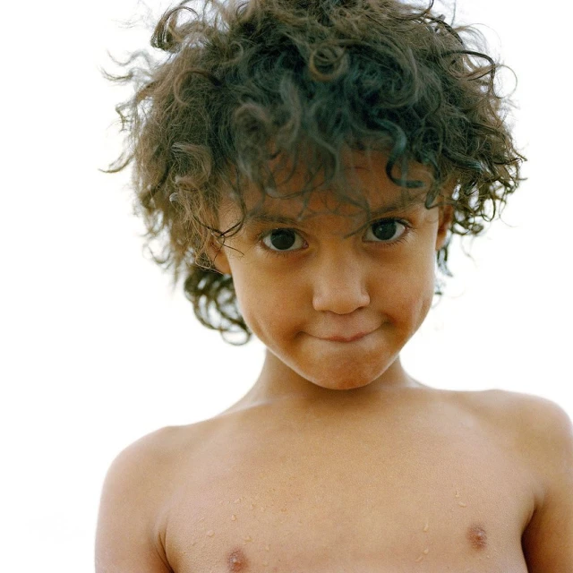 Close-up portrait of a smiling child with curly brown hair against a bright white background, showing their upper torso.