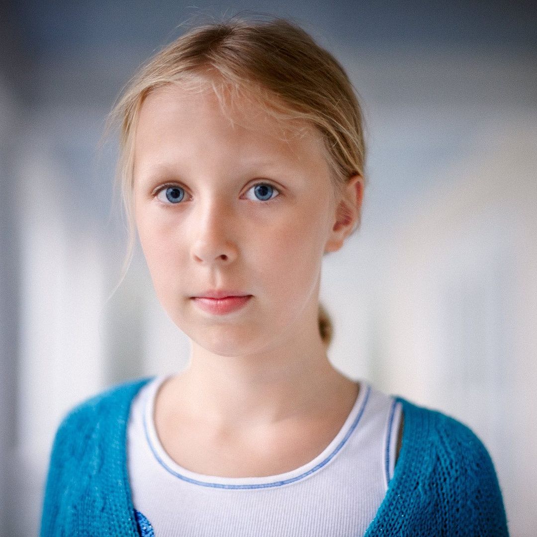 Portrait of a blond child with blue eyes looking directly at the camera, wearing a blue cardigan and white top in an indoor setting.