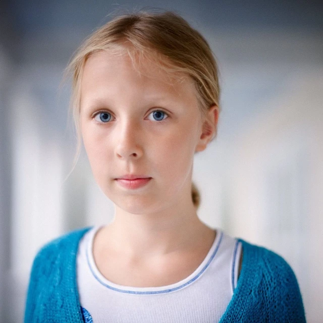 Portrait of a blond child with blue eyes looking directly at the camera, wearing a blue cardigan and white top in an indoor setting.