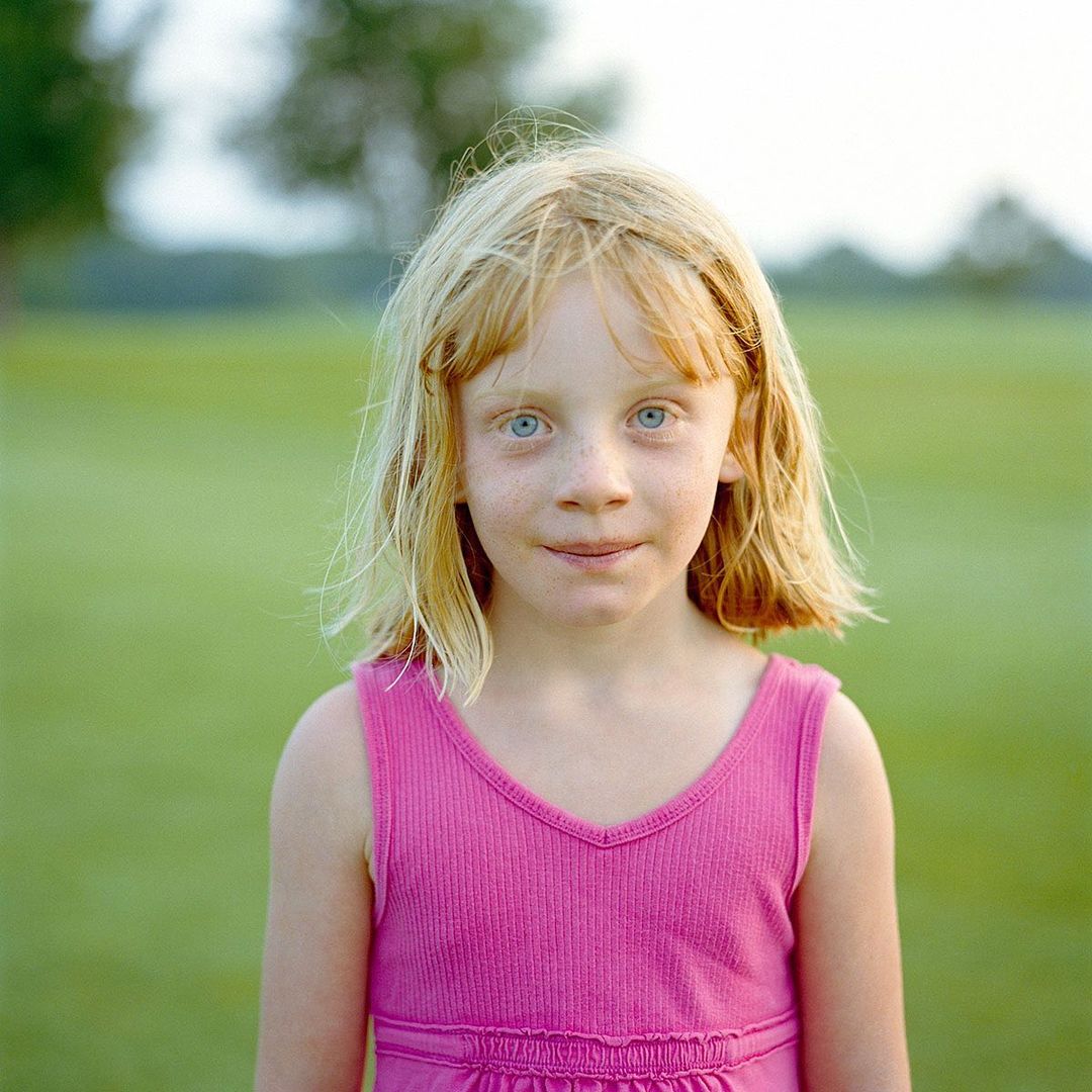 A young child with light blonde hair and blue eyes poses in a pink shirt, standing in a grassy outdoor field with trees in the background.