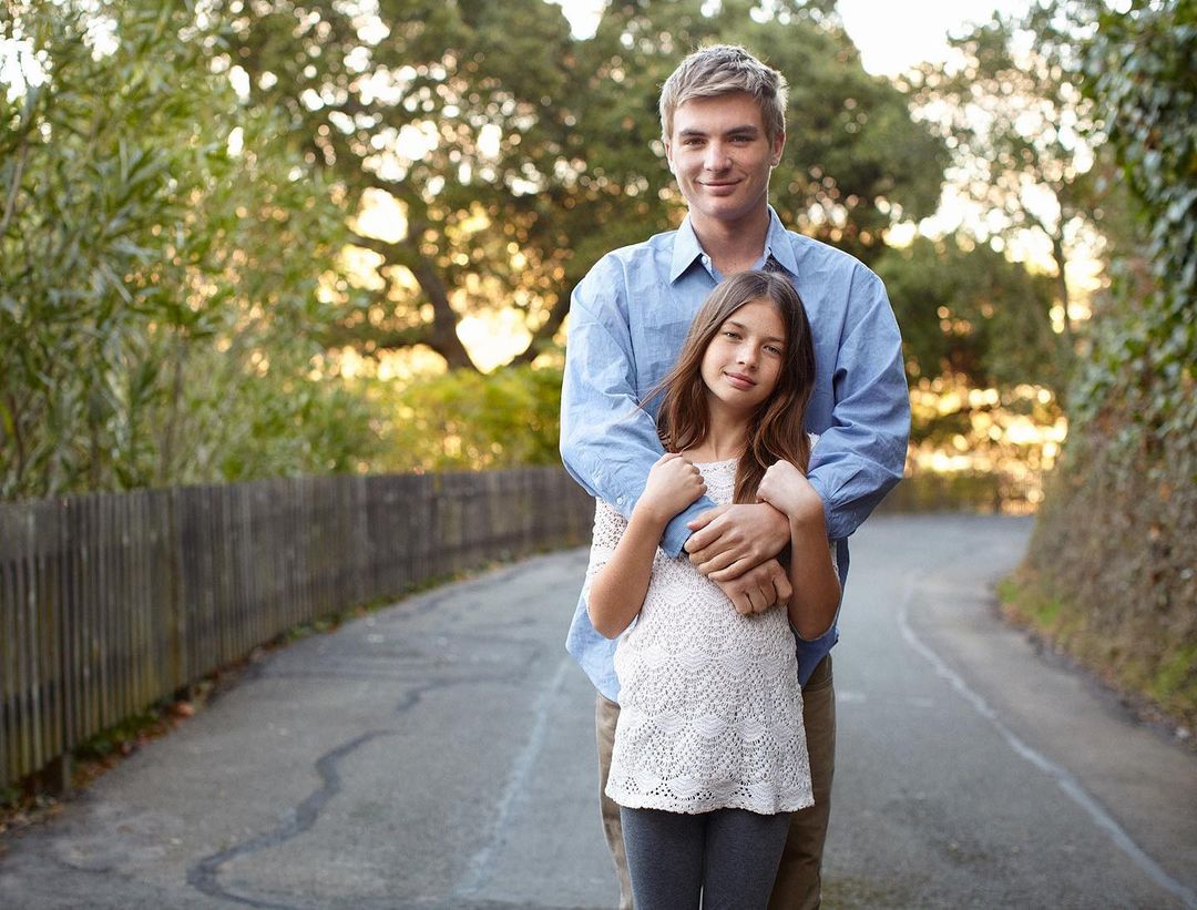Father and daughter pose for a portrait on a path outdoors, surrounded by trees and greenery, creating a warm and family-oriented scene.