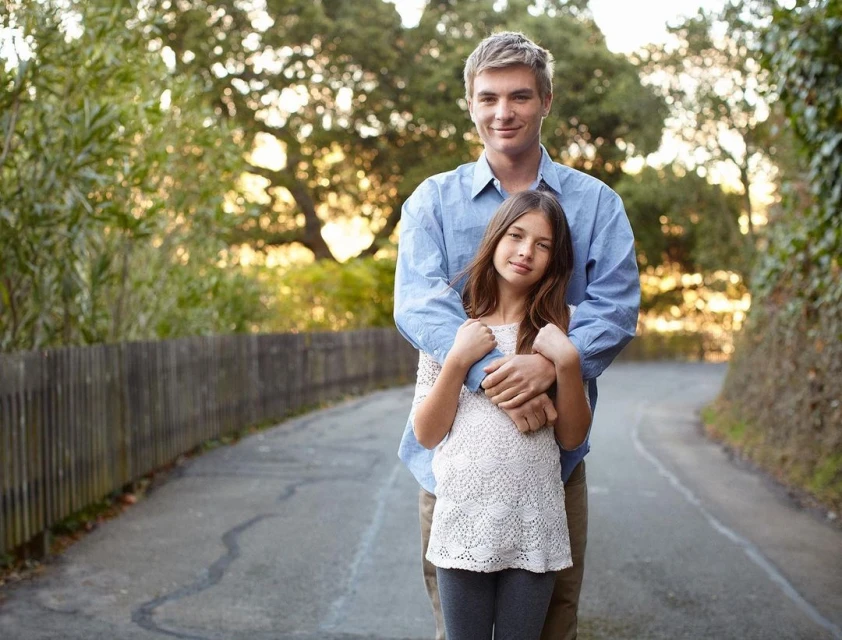Father and daughter pose for a portrait on a path outdoors, surrounded by trees and greenery, creating a warm and family-oriented scene.
