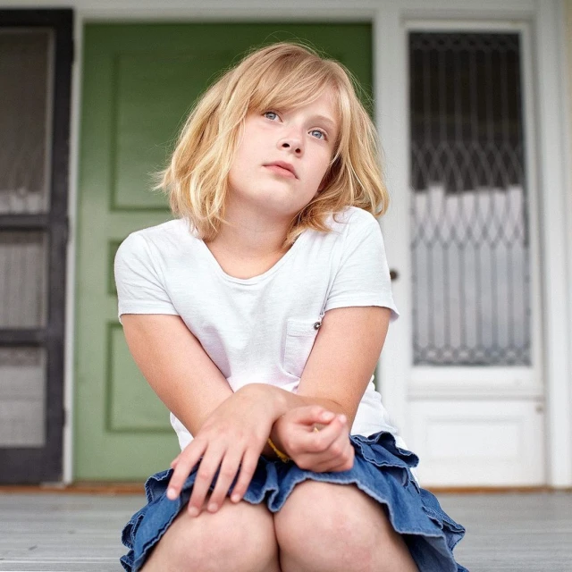 Blonde girl in white t-shirt and denim skirt sits on a porch with green door in the background, looking upwards.