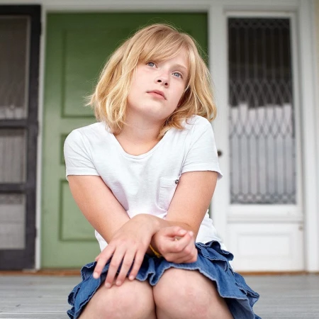 Blonde girl in white t-shirt and denim skirt sits on a porch with green door in the background, looking upwards.