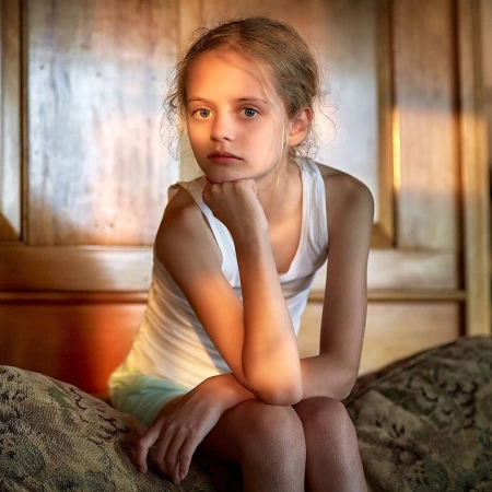 Portrait of a young fair-skinned girl with blonde hair, wearing a white tank top, sitting on a patterned couch in a warm indoor setting.