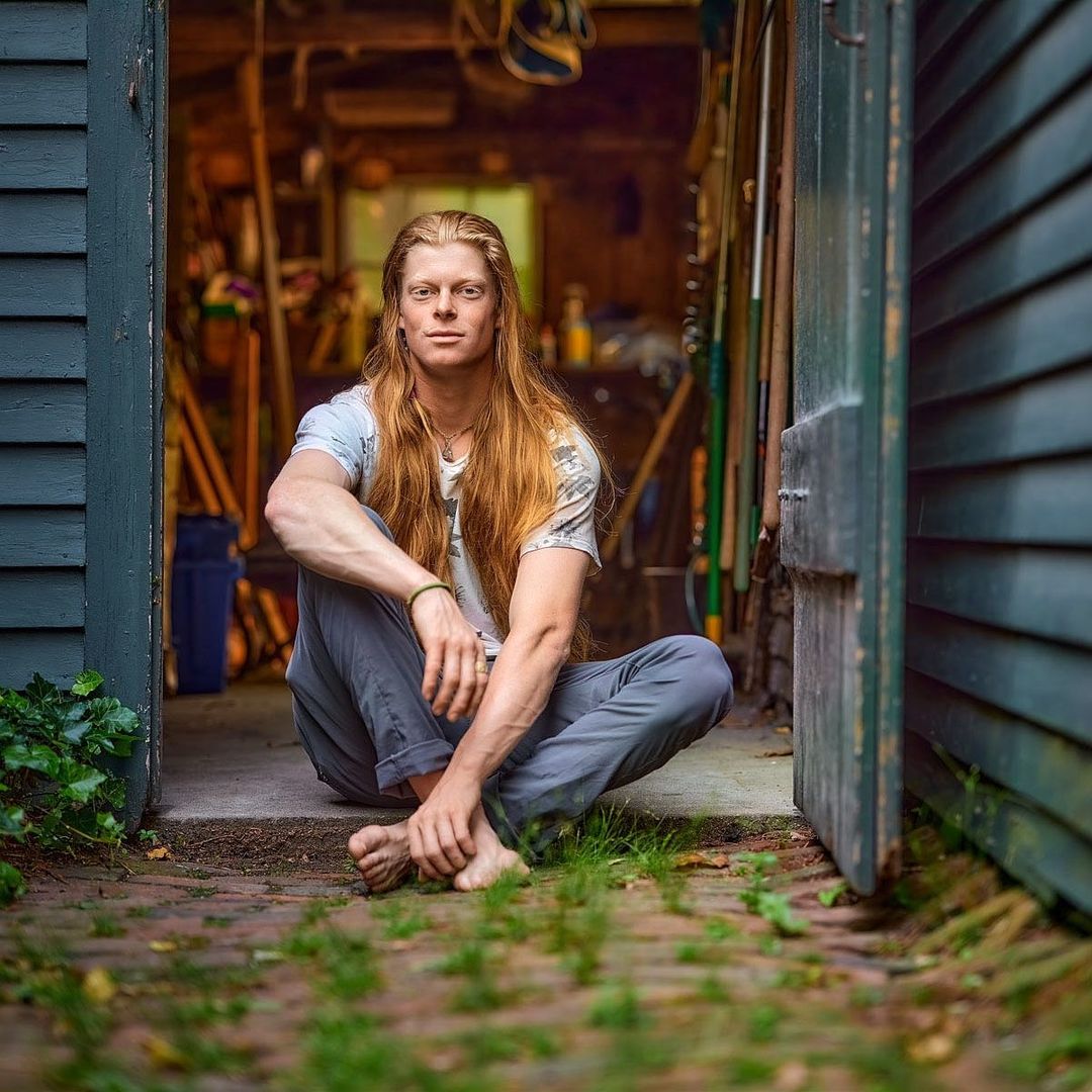Adult male with long hair sits barefoot in the doorway of a shed, wearing casual clothing. The scene is naturally lit outdoors.