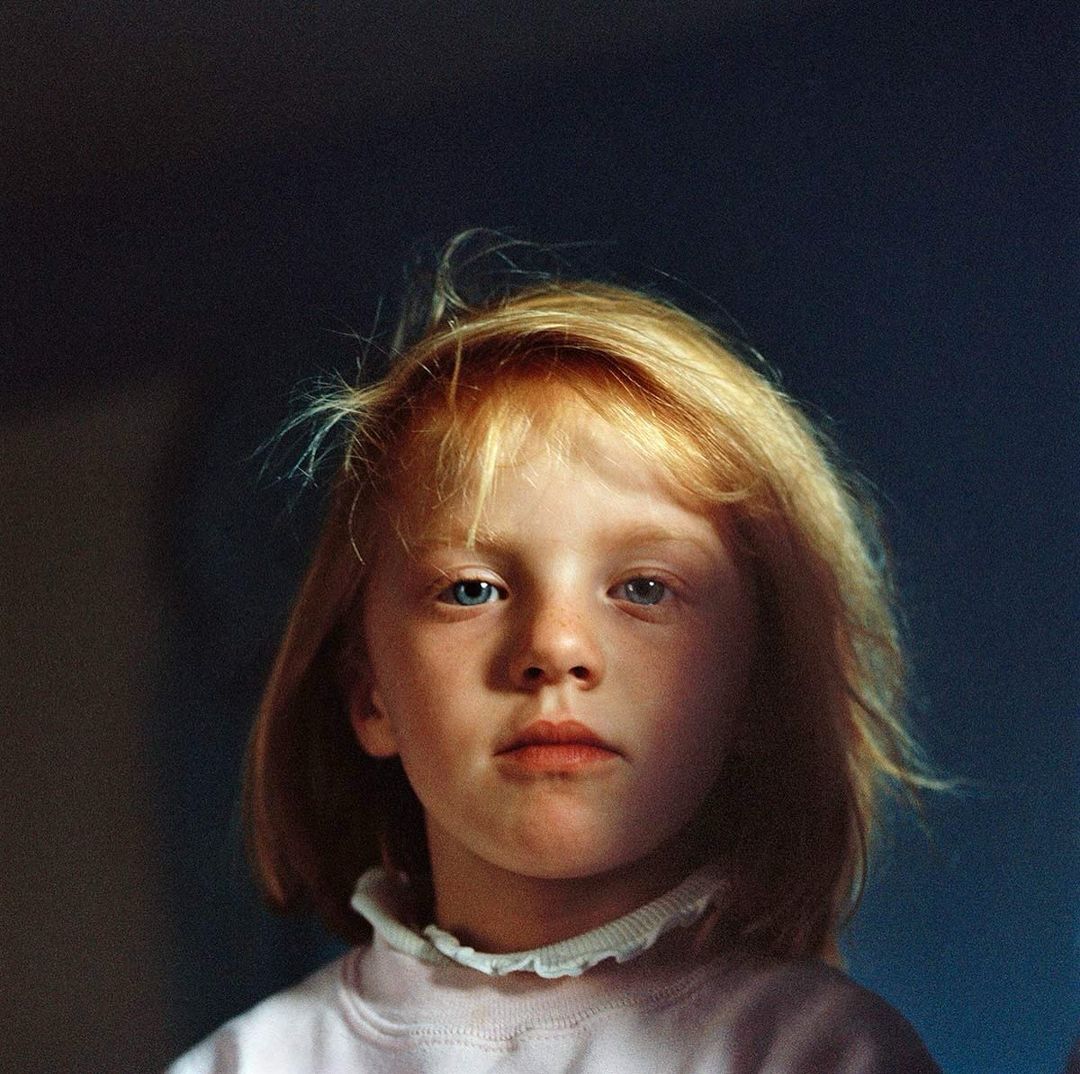 Close-up portrait of a fair-skinned, blond child with blue eyes, wearing a pink sweater with a ruffled collar, set against a dark background.