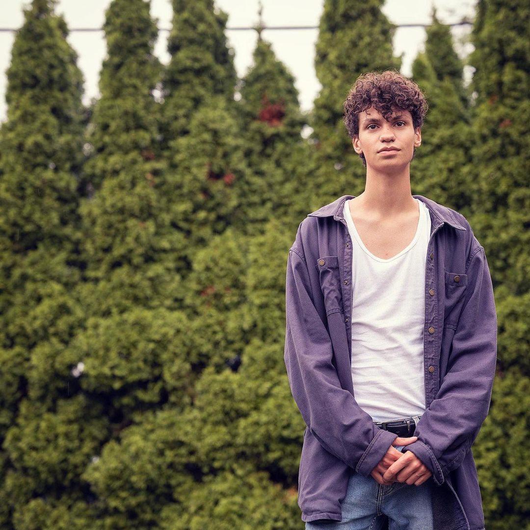 Portrait of a young adult male with curly hair, wearing a white t-shirt and purple shirt, posing outdoors in front of evergreen trees.