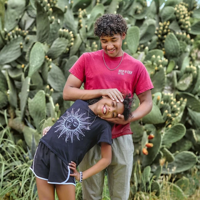 A joyful image of a teen and child in front of a cactus, the child leaning happily with closed eyes against the smiling teen.